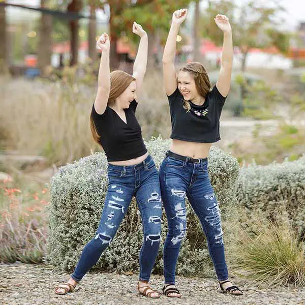 Two high school senior girls in ripped jeans smiling and cheering together outdoors.