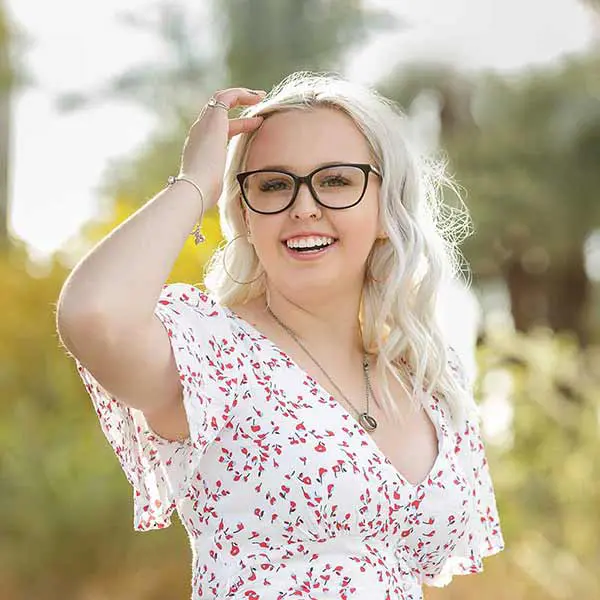 Senior girl with blonde hair and glasses smiling in a floral dress during outdoor session.