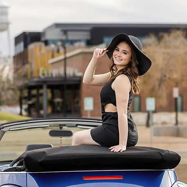 High school senior girl in black dress and wide-brim hat posing on a convertible during her Mesa senior photo shoot.