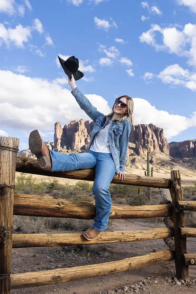 College senior girl smiling in denim outfit, sitting on a wooden fence in front of the Superstition Mountains during her senior portrait session.