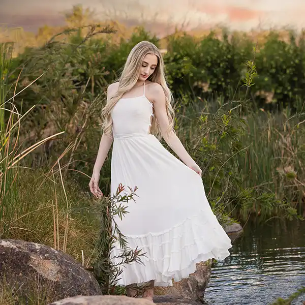 Teen girl in a long white dress standing by the water during her senior portrait session in East Mesa, Arizona.