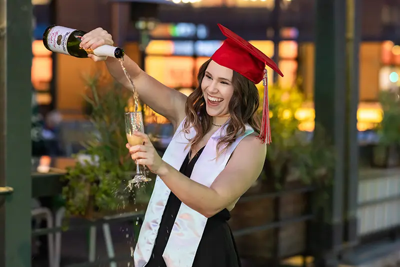 Arizona college graduate in a red cap and gown pouring sparkling cider during her senior photo session.