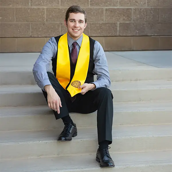College graduate in formal attire and yellow stole sitting on steps and smiling.