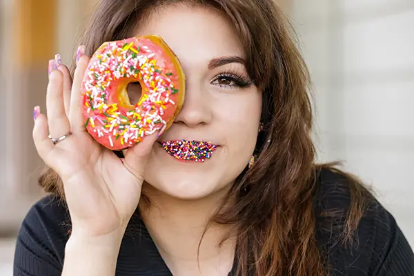High school senior girl with colorful sprinkles on her lips holding a pink frosted donut in front of her eye.