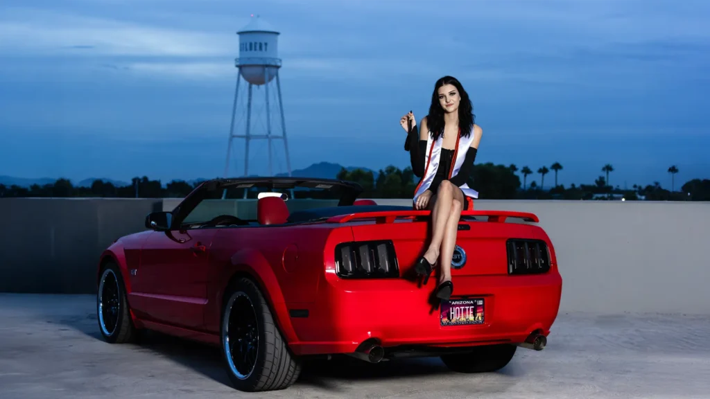 High school graduate sitting on a red Mustang at night holding her tassel, with the Gilbert water tower in the background during her senior pictures.