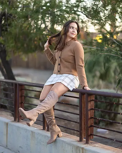 High school senior girl sitting on a railing wearing fall fashion with a tan sweater and boots.