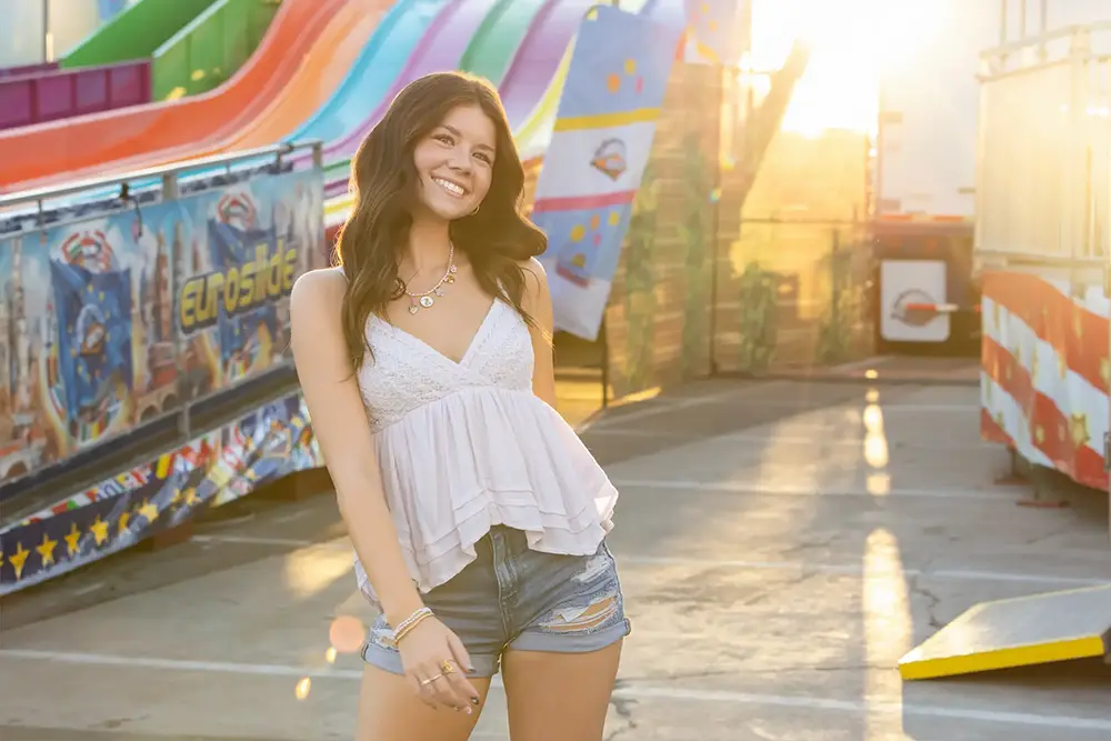 Smiling senior girl in a white top and denim shorts standing near a colorful slide at sunset at the Arizona State Fair.