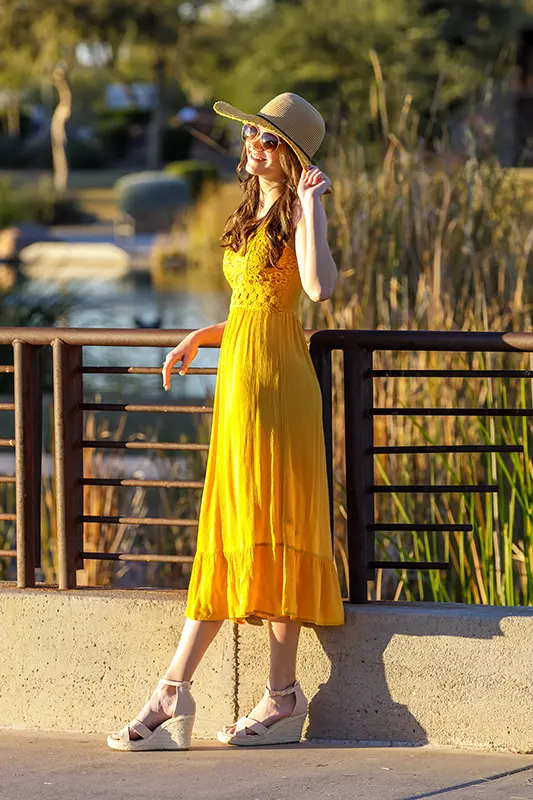 High school senior girl in a yellow dress and sunhat smiling on a bridge during golden hour.