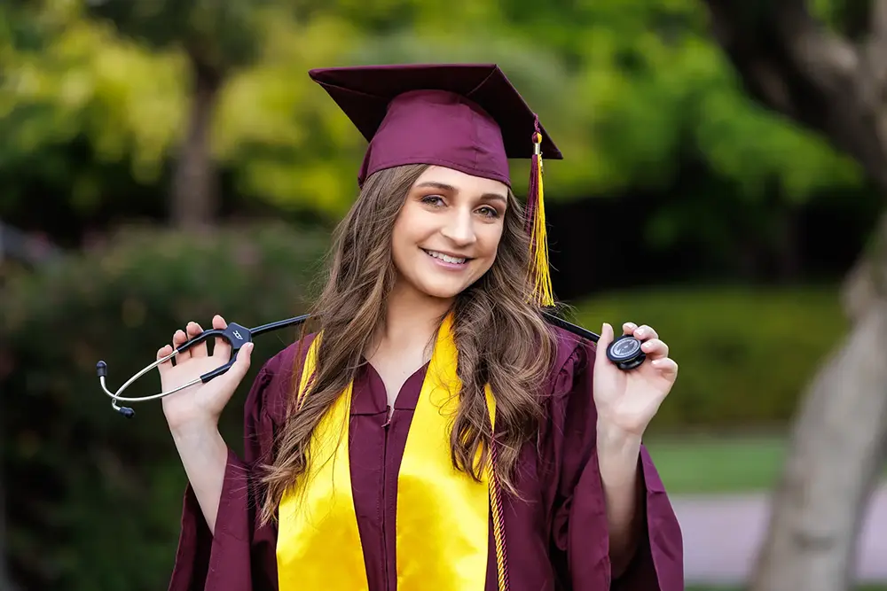 ASU graduate in a maroon cap and gown holding a stethoscope and smiling outdoors.
