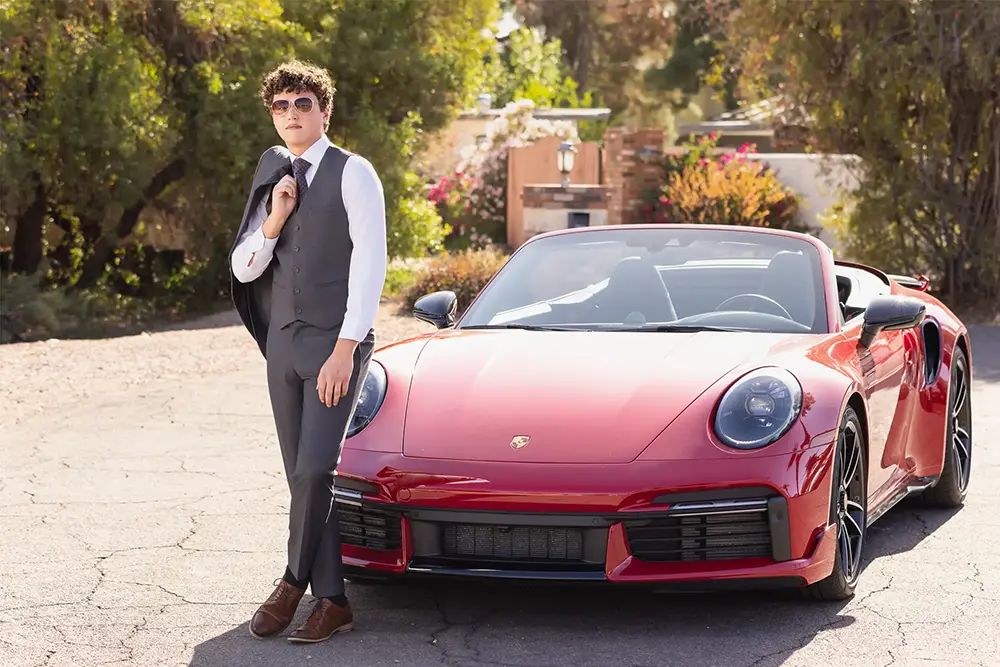 High school senior boy in a sharp gray suit leaning on a red Porsche during his senior portrait session in Chandler, Arizona.