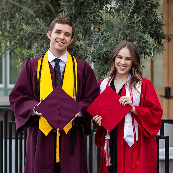 Brother and sister wearing college graduation gowns and holding caps during their portrait session.