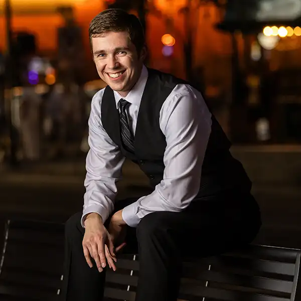 Smiling college senior guy sitting on a bench in formal attire during a nighttime portrait session.