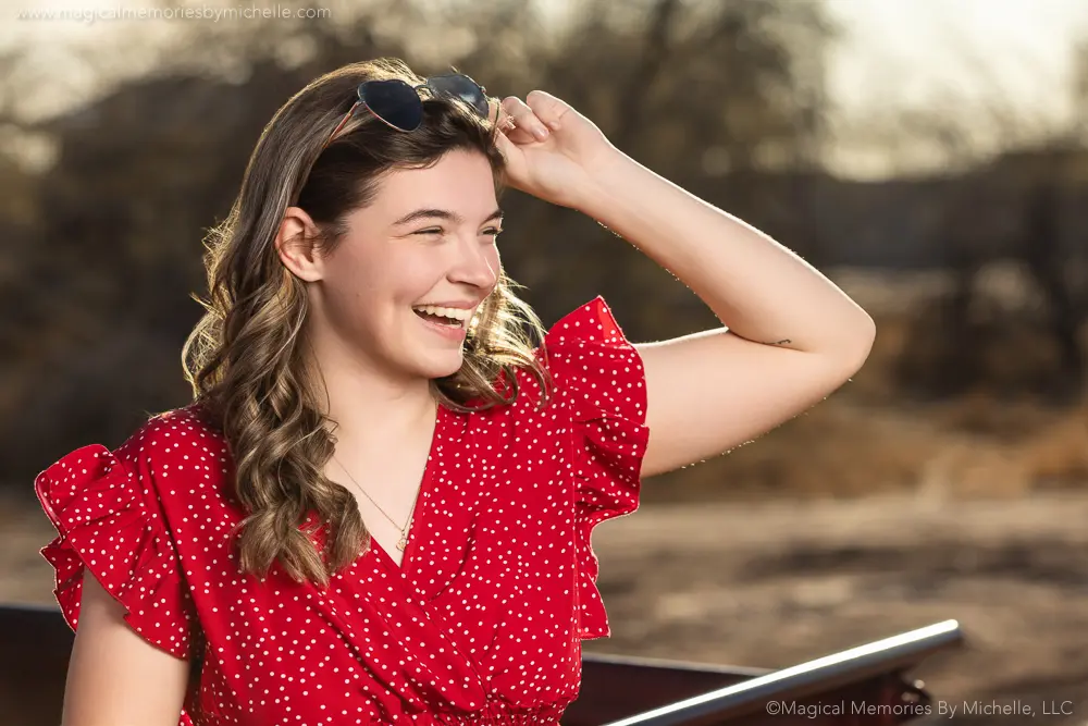 Teen girl wearing a red polka dot top and denim shorts for her senior pictures, posing by a vintage truck with heart sunglasses.