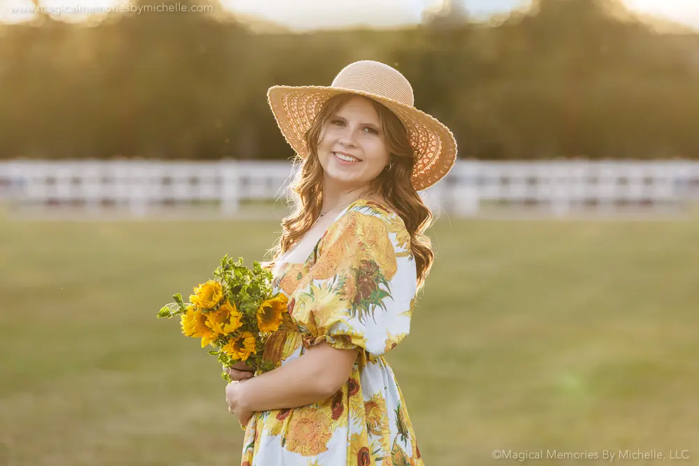 Senior girl in a sunflower-print dress holding a bouquet, wearing a sun hat for a cheerful summer senior photo.