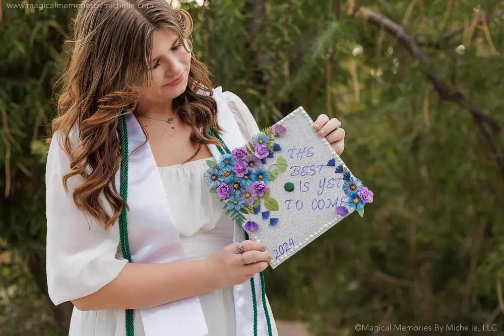 Senior girl in white dress holding a silver graduation cap decorated with colorful flowers and the quote “The best is yet to come.”