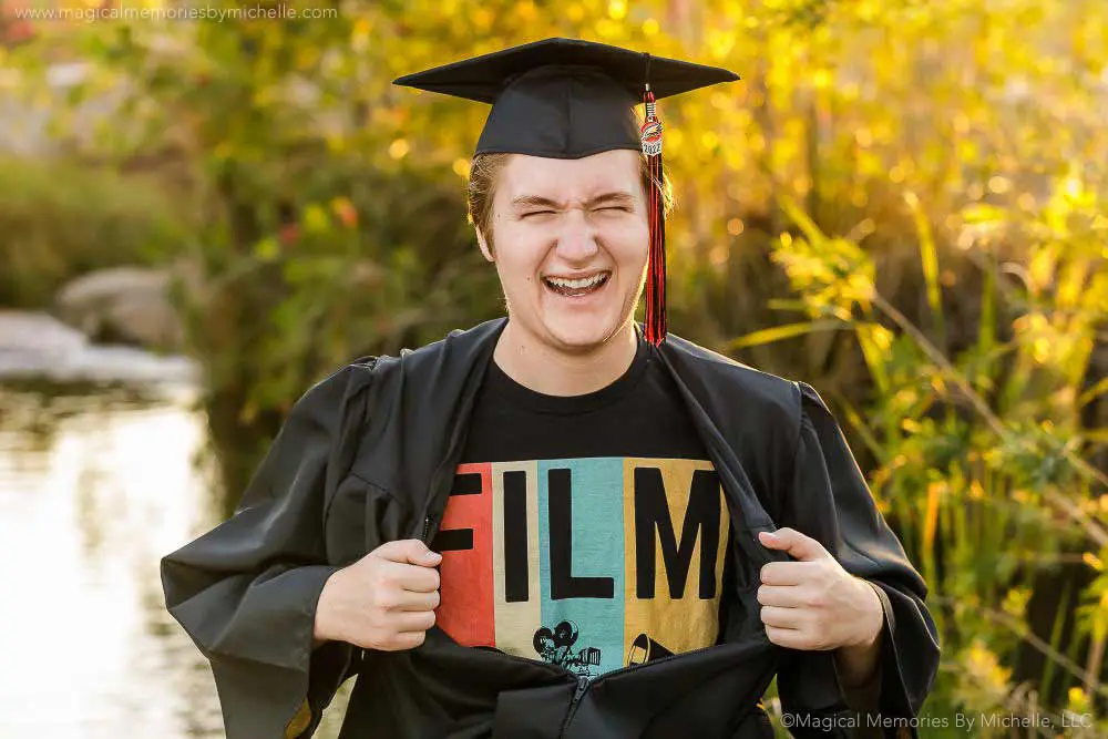 Senior guy wearing a cap and gown laughing while showing his film t-shirt during graduation photos in Mesa, Arizona.