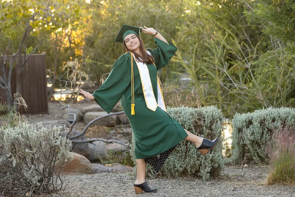 Senior girl posing with a fun kick in her green cap and gown in a nature setting in Mesa, Arizona.