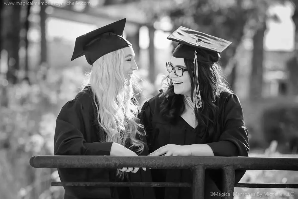 Two senior girls in black caps and gowns smiling at each other during a black and white graduation photo session in Mesa, Arizona.