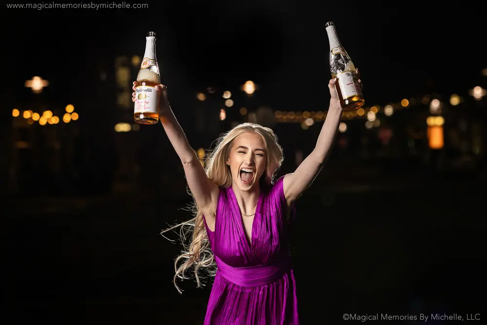 Senior girl in a purple dress holding two bottles of sparkling cider during a fun night senior photo shoot.