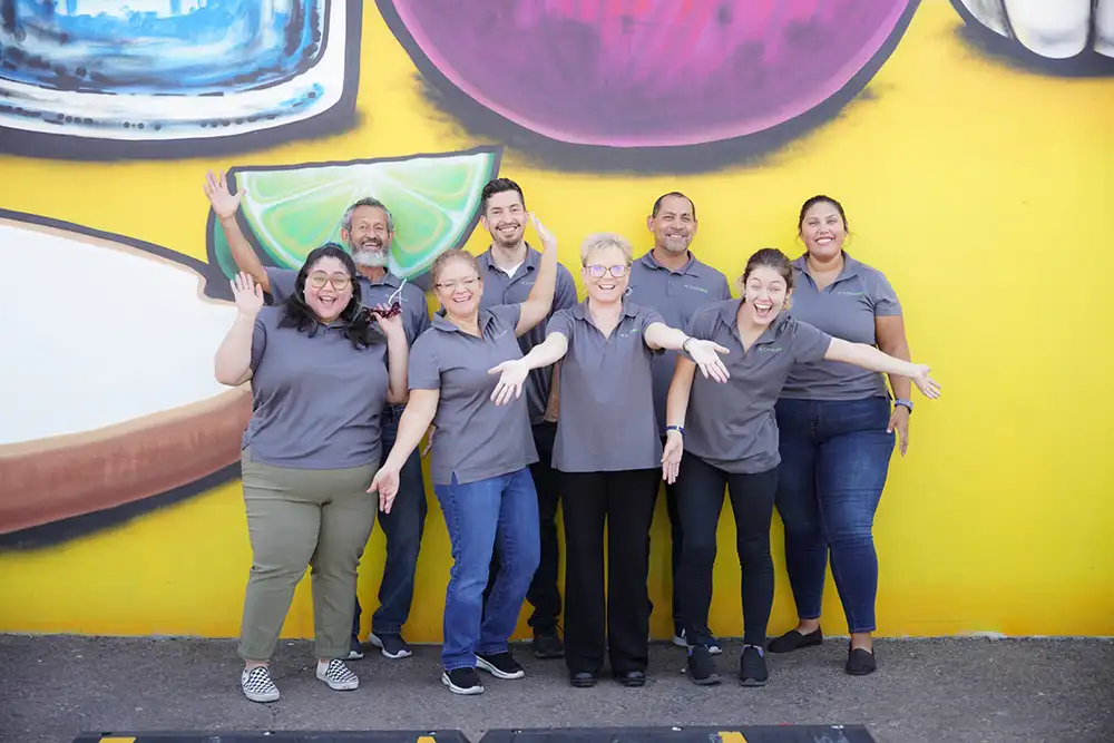 Group of Nourish Phoenix team members smiling and posing in front of a bright yellow wall with fruit artwork.