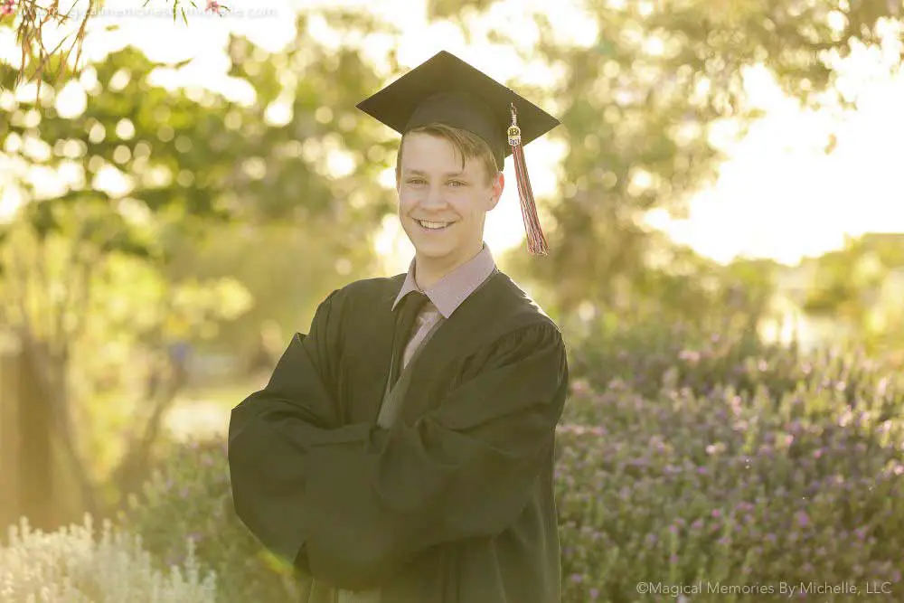 Senior guy in black cap and gown smiling with arms crossed during golden hour graduation pictures in Mesa, Arizona.