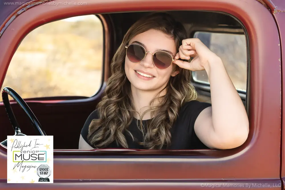Senior girl wearing sunglasses and a black shirt, smiling while leaning on the window of a vintage red truck during her published senior photo session in Mesa, Arizona.