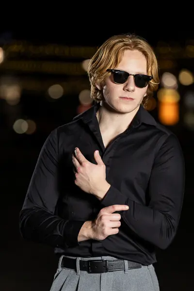 Senior guy wearing sunglasses and a black button-down shirt posing at night with city lights in the background during his Arizona photo session.