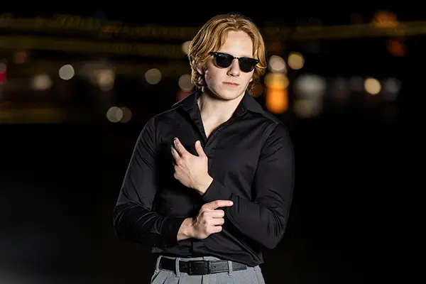 Senior guy wearing sunglasses and a black shirt during a stylish nighttime photo session with city lights in the background.