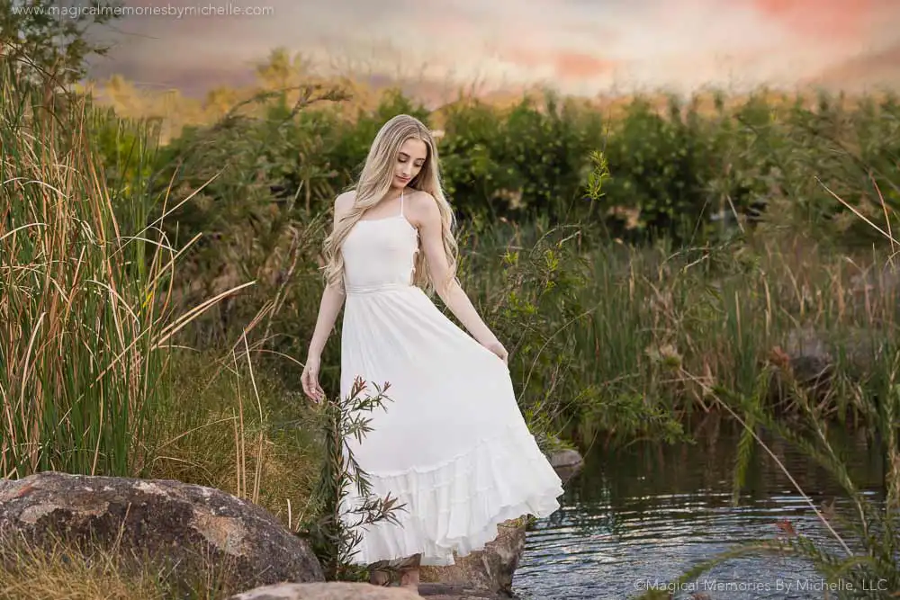Senior girl in a white dress poses by the water at a lush portrait location in East Mesa during sunset.