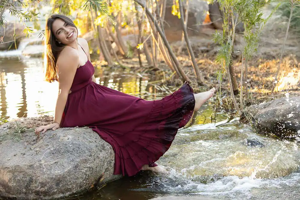 Senior girl in a burgundy dress smiling and splashing water with her foot during a candid senior photo session in Mesa, Arizona.