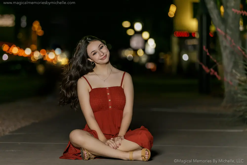 Senior girl sitting on the sidewalk at night in a red dress with glowing bokeh lights in the background.