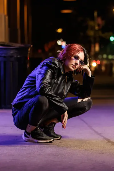 Senior girl in sunglasses and black leather jacket crouching on a city sidewalk at night for her senior portrait session in Arizona.