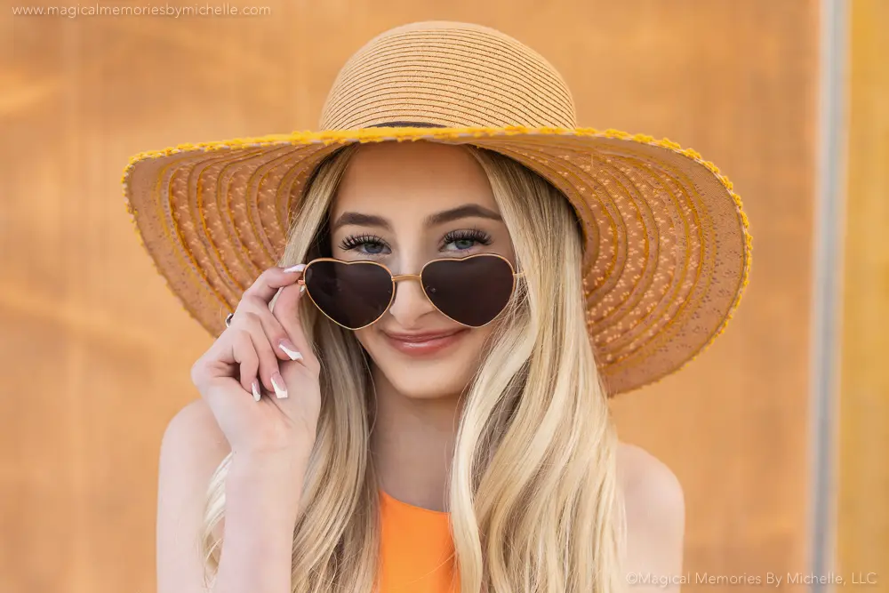 Senior girl wearing heart-shaped sunglasses and a wide-brimmed sun hat during her summer senior pictures in Gilbert, Arizona.