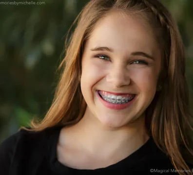A confident teen girl with long brown hair and braces smiles brightly in front of soft greenery.