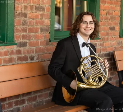 High school senior in a formal suit sitting on a bench with his French horn during senior pictures.