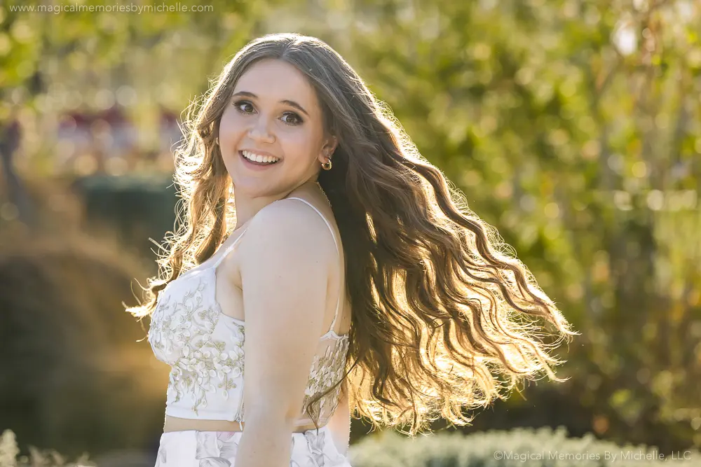 Senior girl with long, highlighted wavy hair in a two-piece white dress, twirling outdoors in golden light.