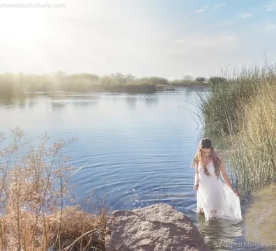 Senior girl in white boho dress walking in lake water with sun flare during Chandler senior photo session.