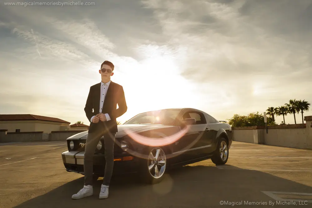 Senior boy wearing a tuxedo stands confidently in front of a black Mustang during a sunset photo session in Arizona.