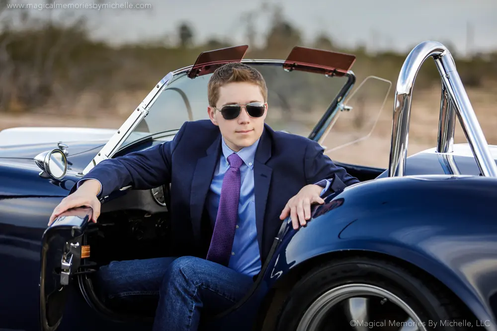 Teen boy in a suit and sunglasses posing in a classic blue convertible for senior pictures in the Arizona desert.