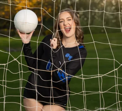 Senior girl smiling through the net while holding a volleyball during her photo session.
