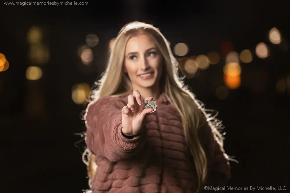 Close-up of a graduation charm held toward the camera, with the senior girl softly out of focus and bokeh lights in the background.
