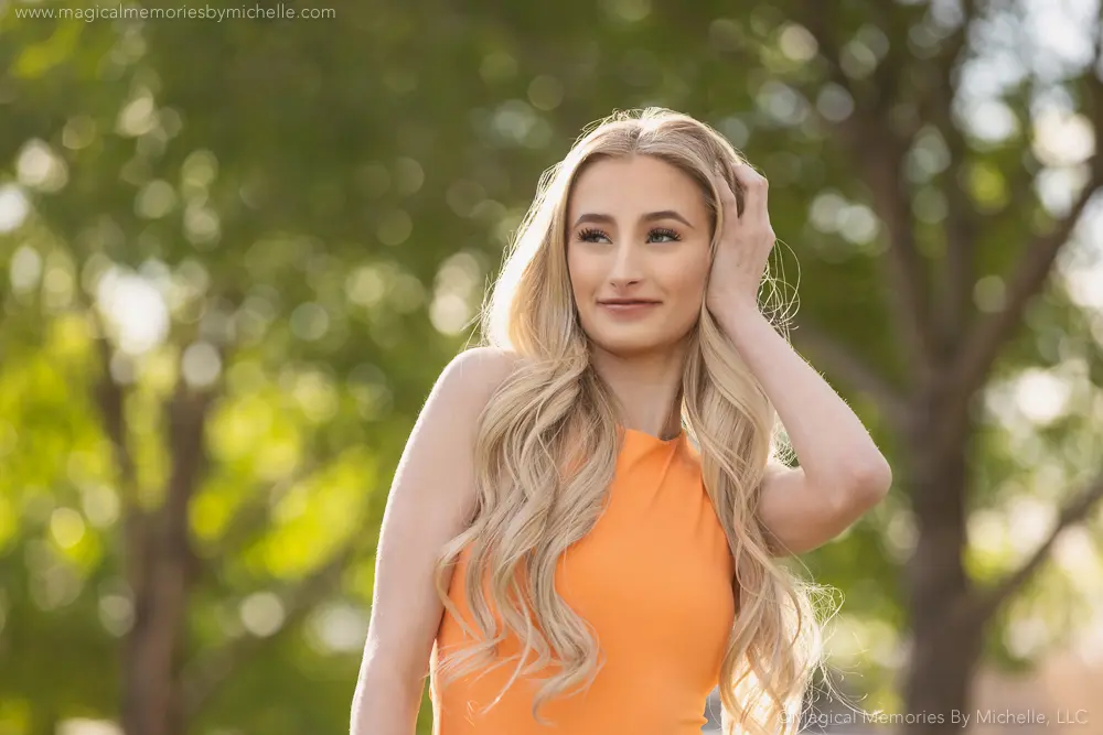 Smiling senior girl in an orange dress stands outdoors with soft bokeh and trees in the background.