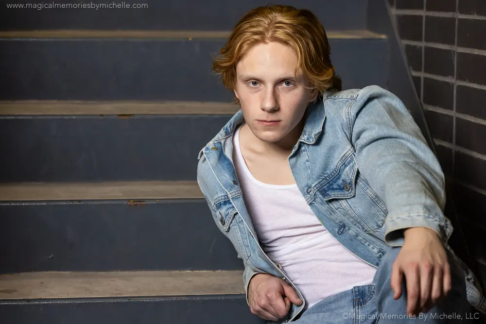 Senior guy wearing a denim jacket and white tee, seated on stairs in an urban setting for a confident and cool senior portrait.