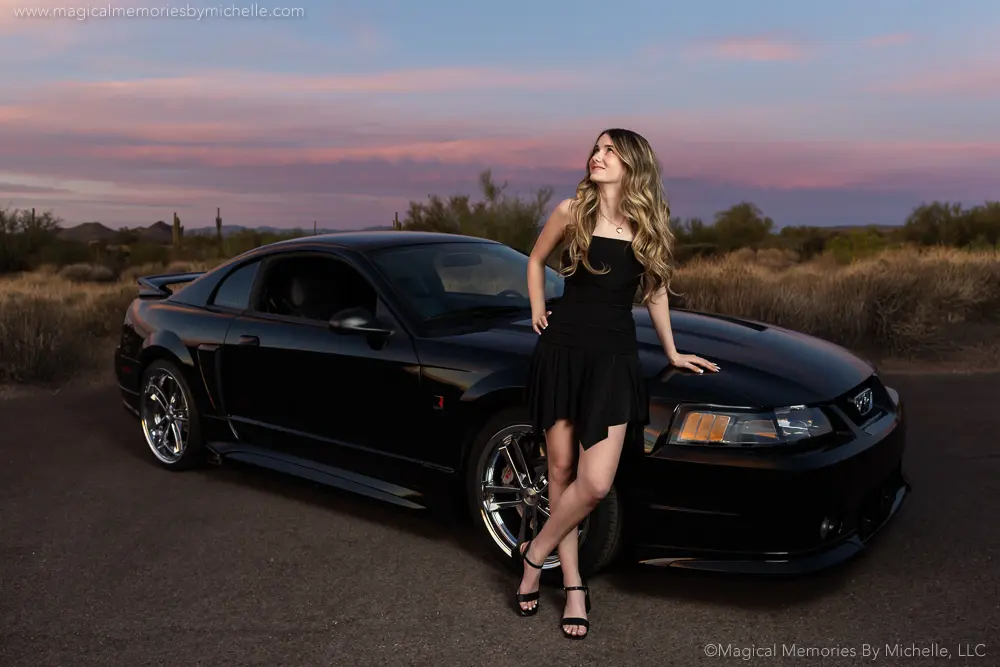 High school senior girl in a black dress posing with a black Mustang during sunset in the Arizona desert.