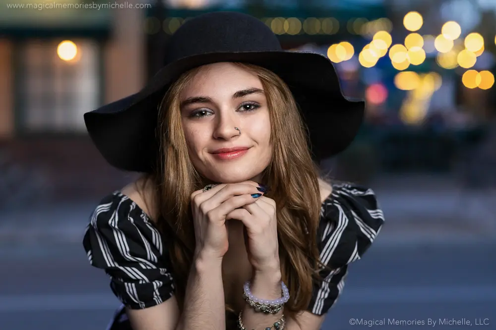 Teen girl wearing a wide-brim hat and natural makeup, smiling during a nighttime senior photo shoot in Arizona with city lights in the background.