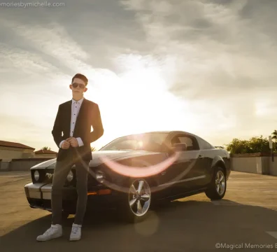 Teen in a black suit poses with a black Mustang on a rooftop parking garage in Mesa at sunset.