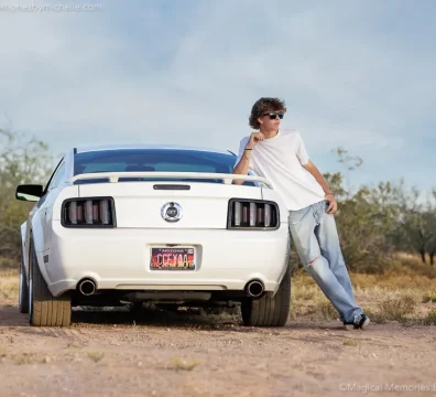 Senior boy in white t‑shirt and jeans leaning on a Mustang during desert photo shoot.