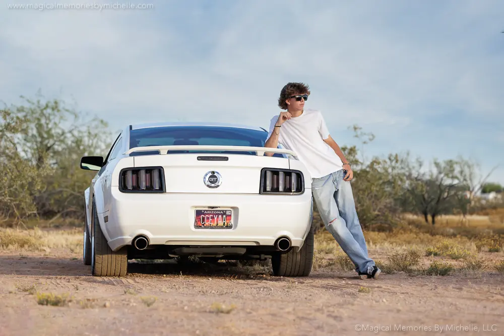 Senior boy in white t‑shirt and jeans leaning on a Mustang during desert photo shoot.