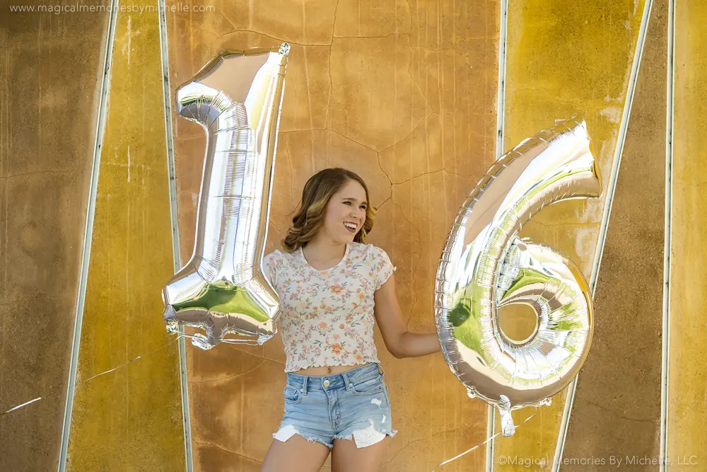 Teen girl in floral top and denim shorts holds silver 1 and 6 balloons for her Sweet 16 birthday photo shoot in Arizona.