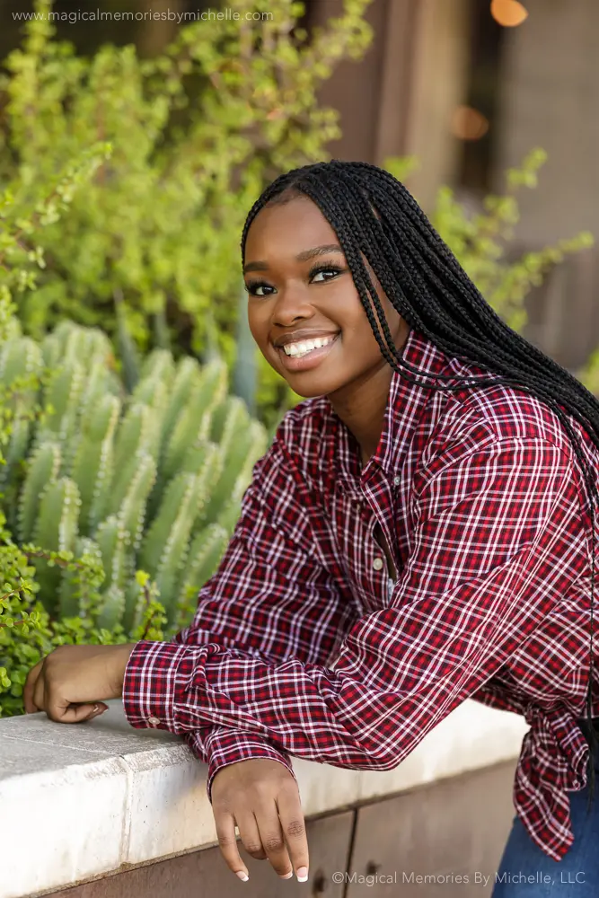 Black high school senior girl with long braids smiling while leaning against a wall with greenery in the background during an outdoor portrait session in Arizona.